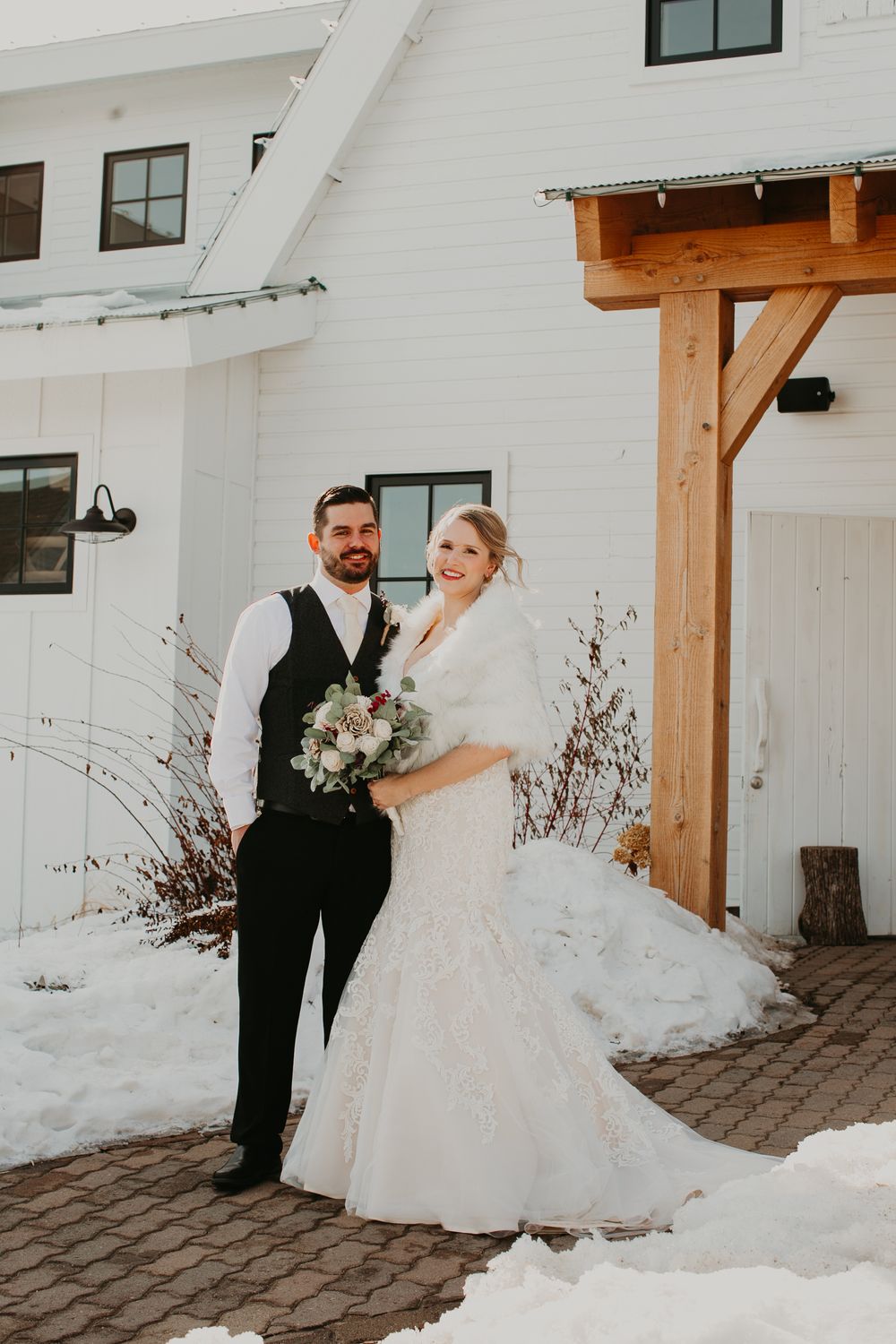 Bride and groom pose together outside a white barn venue in winter, bride holding a bouquet in a lace mermaid gown.