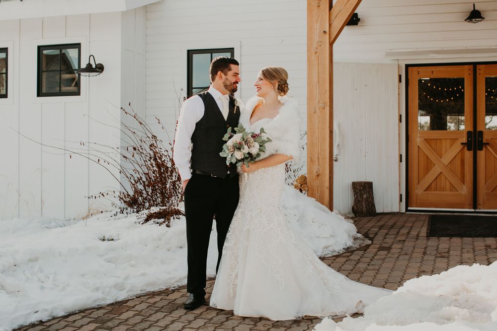 Bride and groom share a romantic gaze outside a white barn wedding venue surrounded by snow and winter scenery.