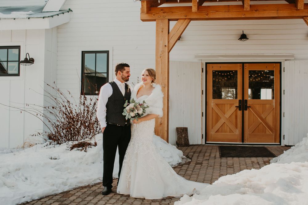 Bride holding bouquet and groom face each other lovingly outside a white barn venue with wooden doors in winter.