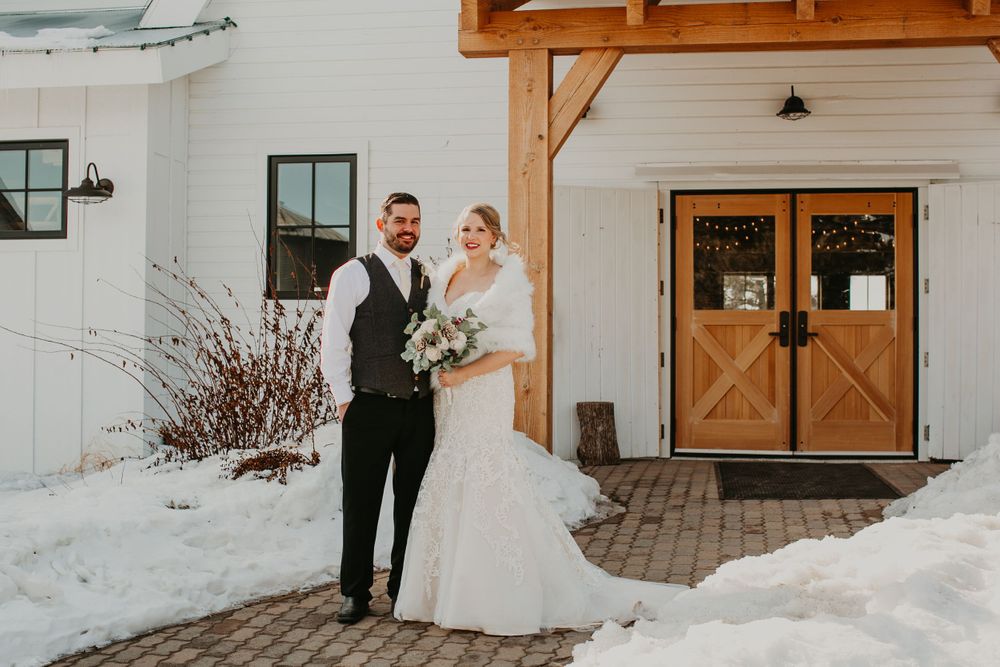 Bride and groom stand together smiling outside a white barn wedding venue with wooden doors in a snowy landscape.