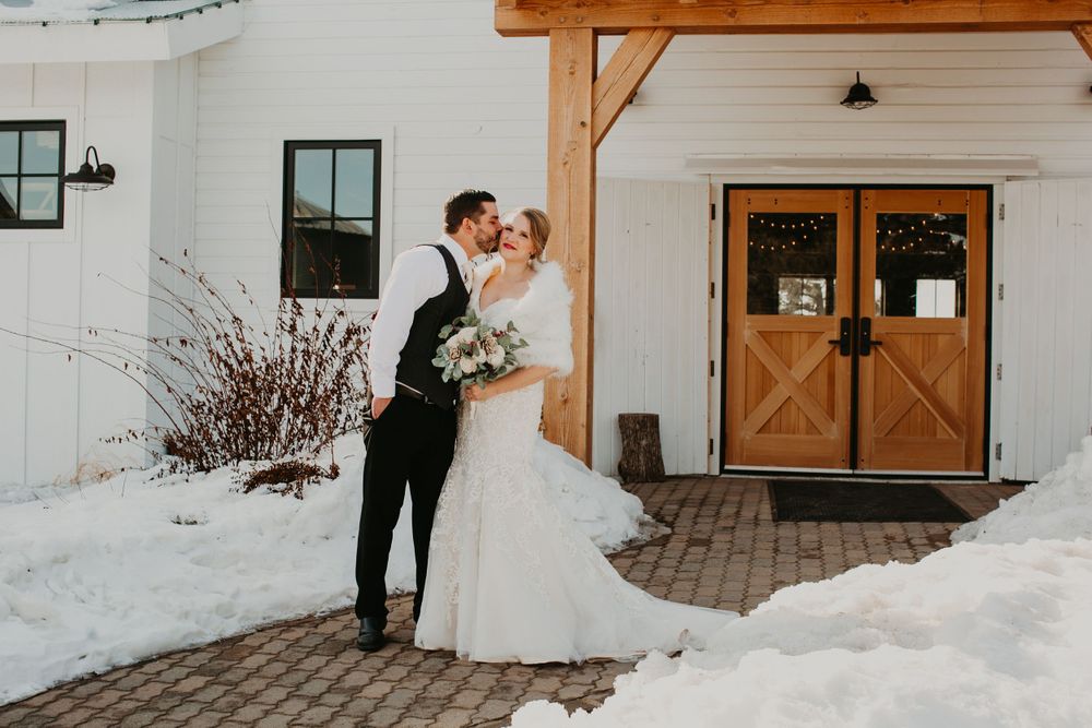 Groom kisses bride outside a white barn wedding venue with wooden doors surrounded by snow in a winter wedding.
