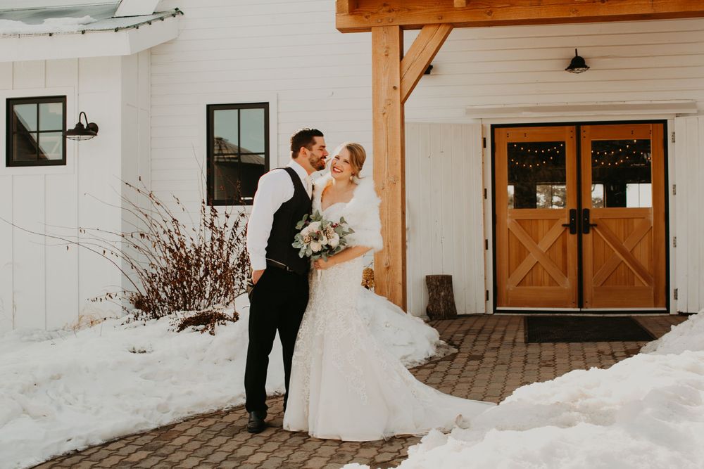 Bride and groom share a romantic kiss outside a white barn wedding venue with wooden doors in a snowy setting.