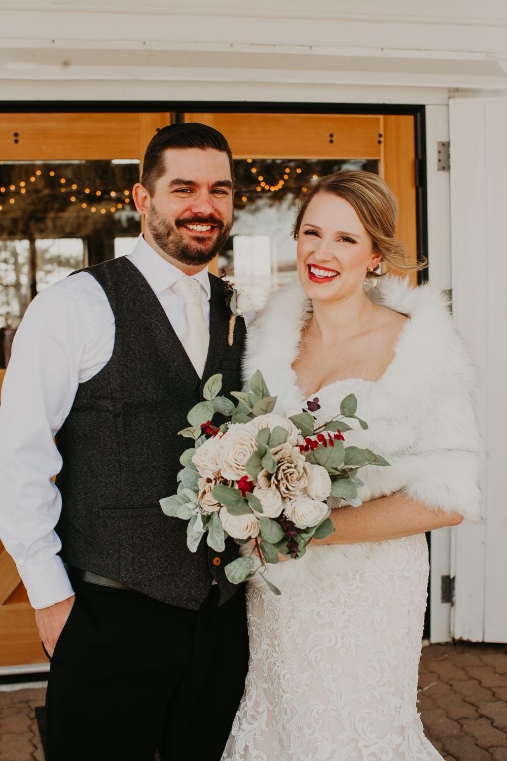 Smiling bride and groom pose together outdoors in winter, bride wearing fur wrap and holding bouquet of roses and berries.