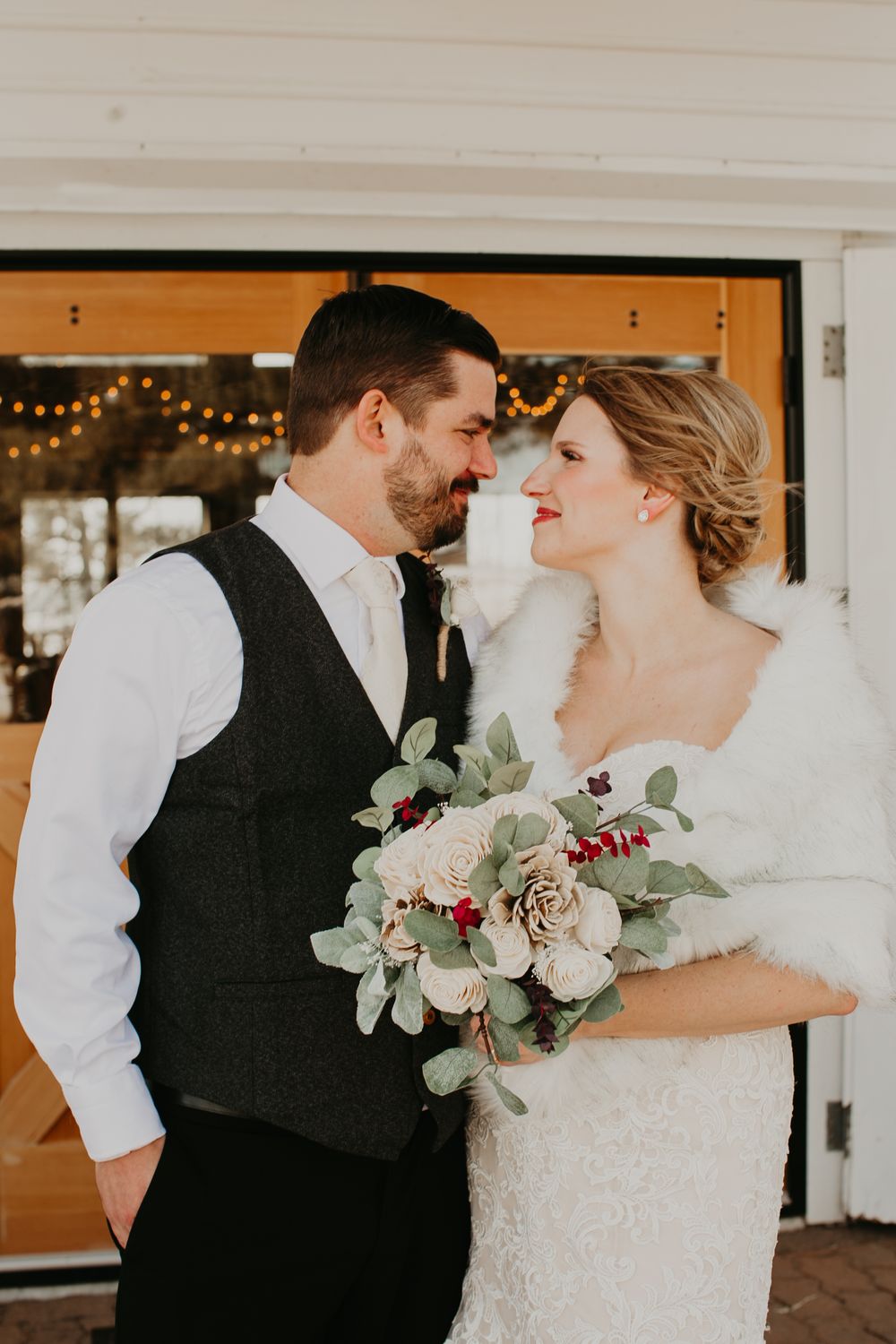Bride in fur wrap and groom in dark vest share an intimate moment holding a winter bouquet of roses and berries.