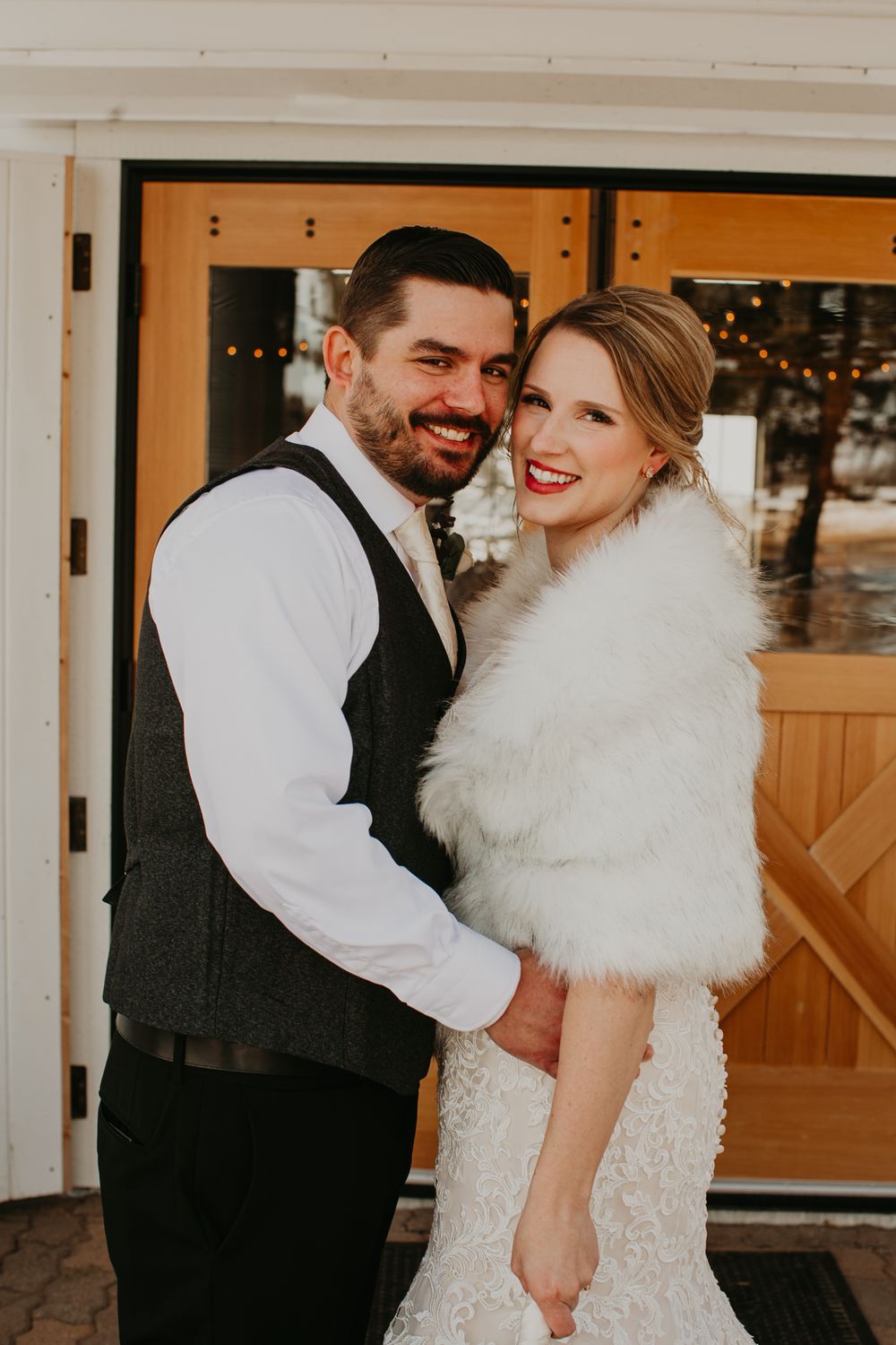 Smiling bride in white fur stole and groom in dark vest embrace warmly in front of wooden barn doors.