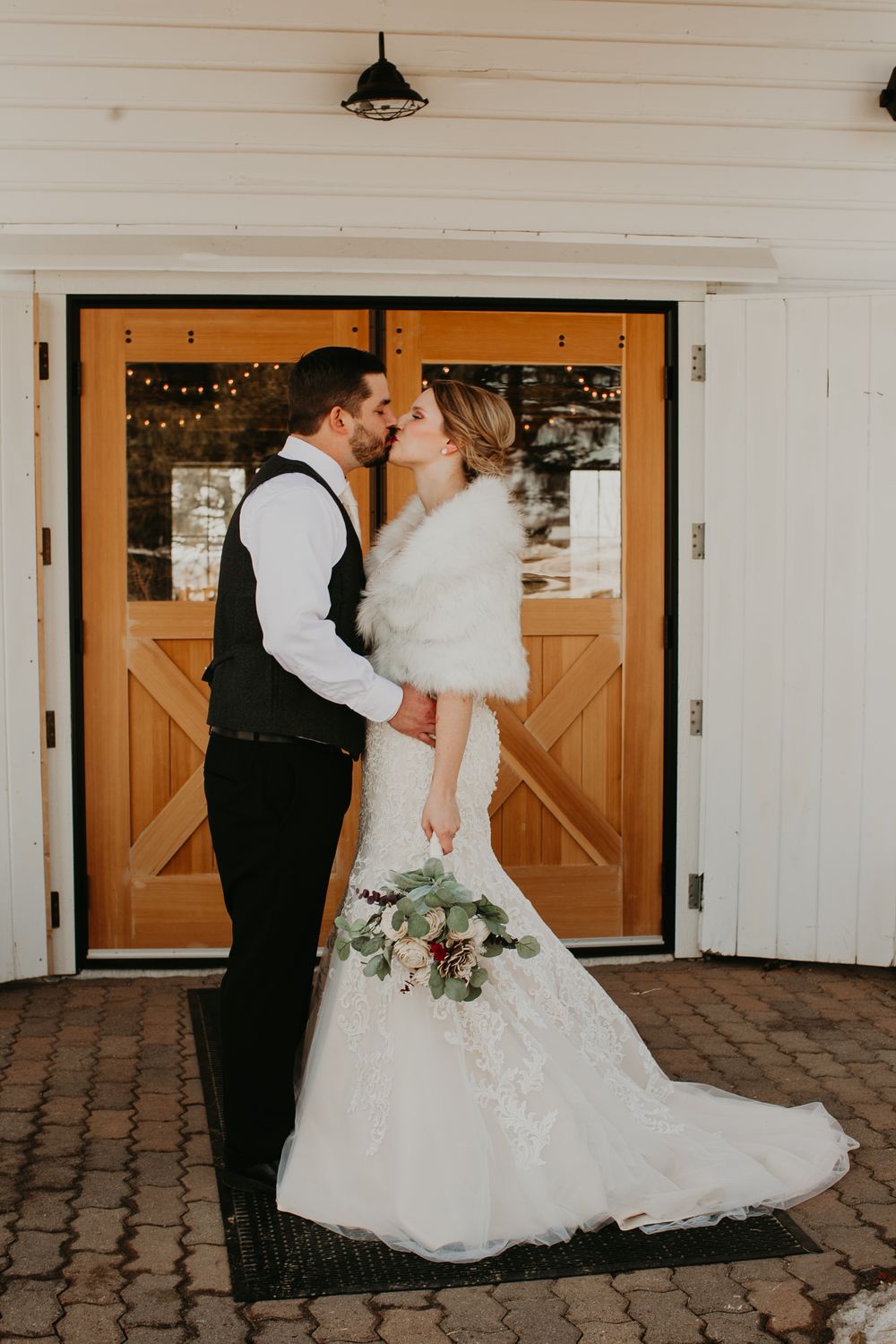 Bride and groom share a romantic kiss in front of rustic wooden barn doors with string lights at winter wedding.