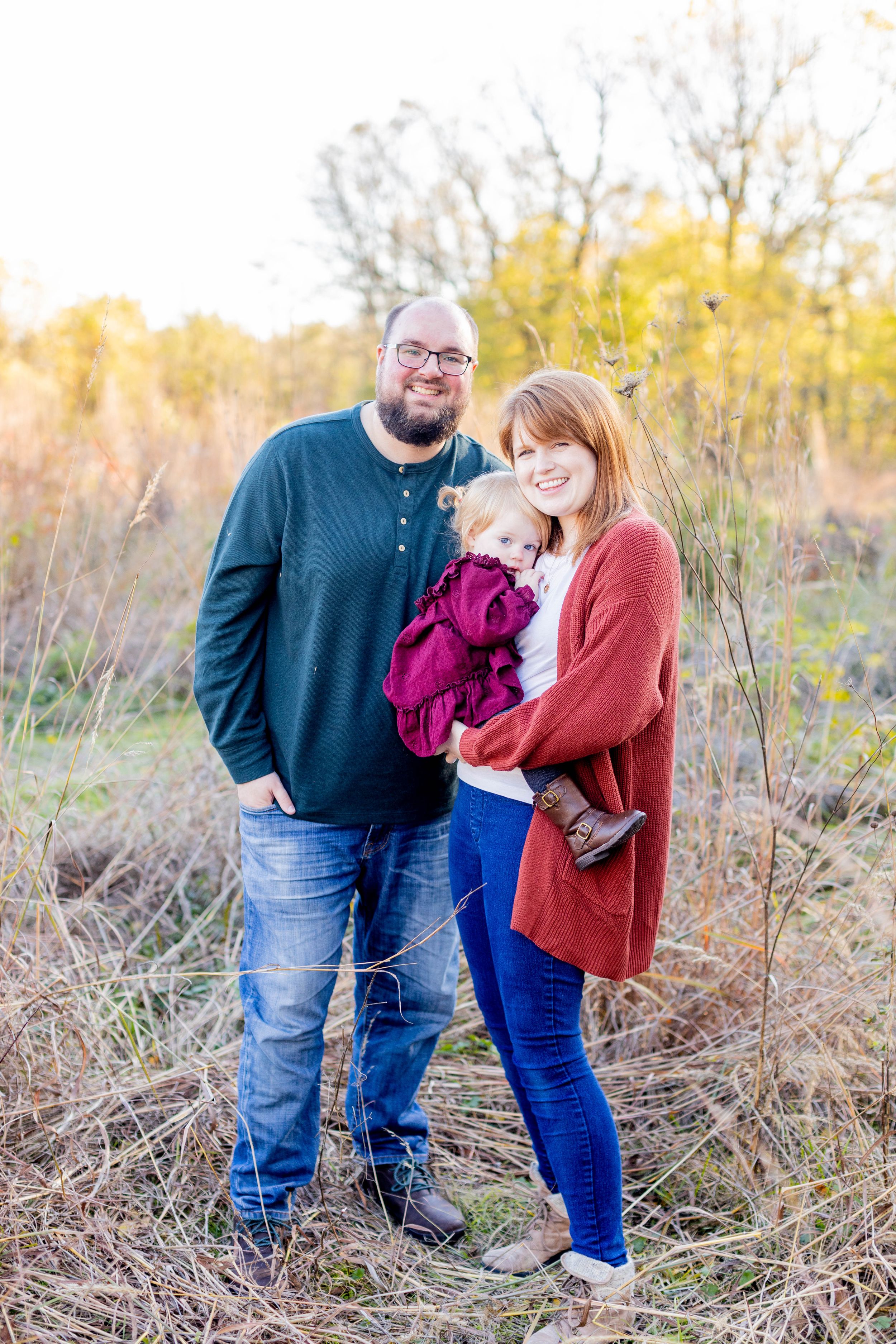 Family outdoor portrait with toddler in burgundy dress surrounded by golden fall foliage in Springfield, MO.
