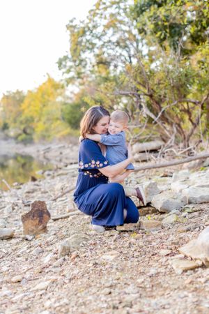 Parent and child embrace while kneeling on rocky shoreline with water and trees in background in Springfield, MO.