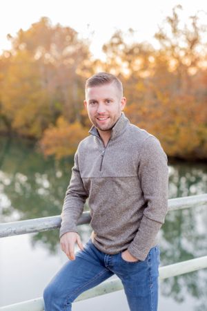Man in grey sweater poses casually on bridge railing with autumn foliage in background in Springfield, MO.