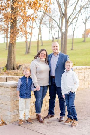 A family of four poses outdoors on a stone path during autumn with fall foliage in the background in Springfield, MO.