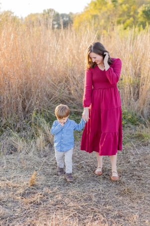 Woman in red dress holding hands with small child in light blue shirt walking through tall grass at sunset in Springfield, MO..