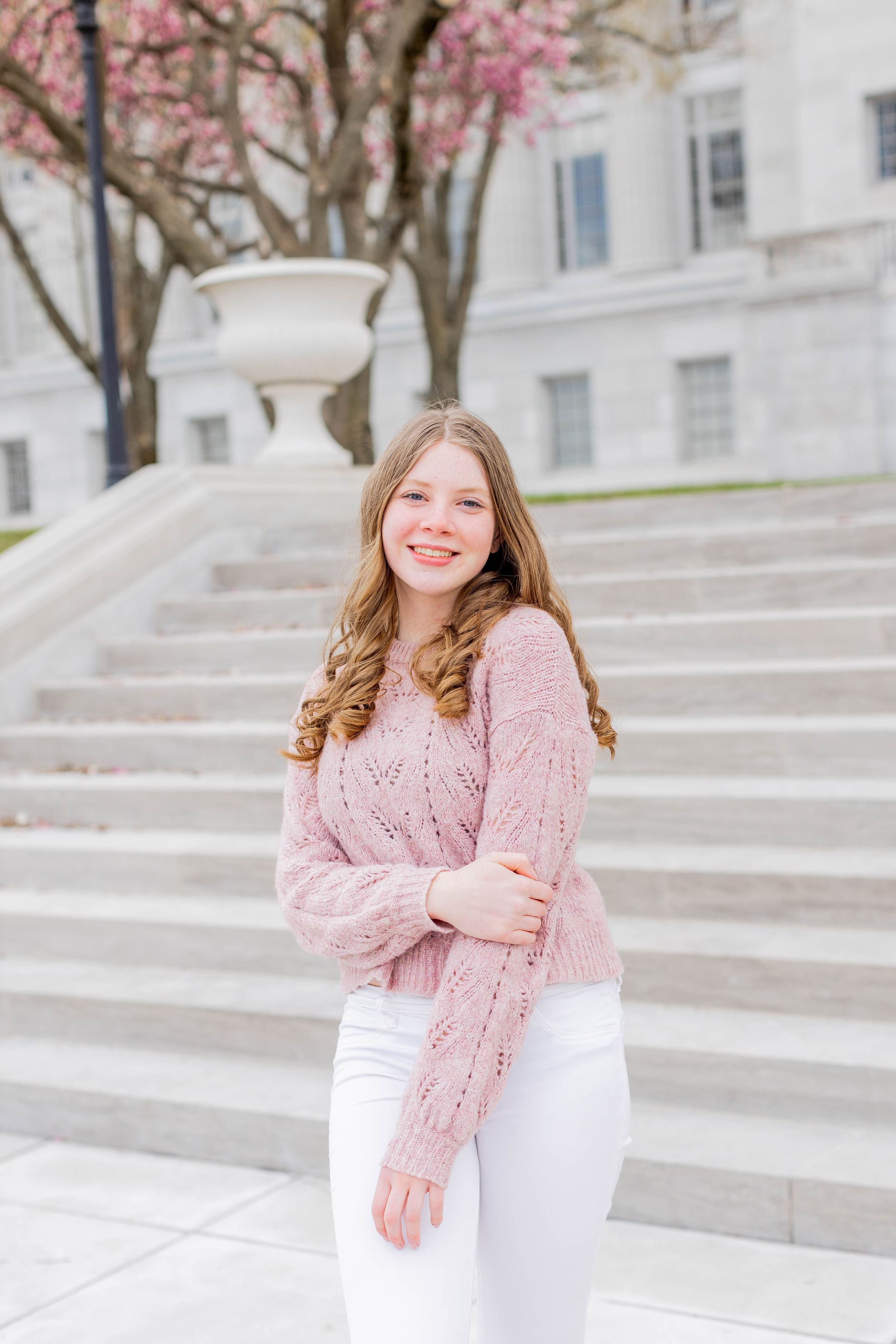 Young woman in pink sweater poses on stone steps with cherry blossoms in background.