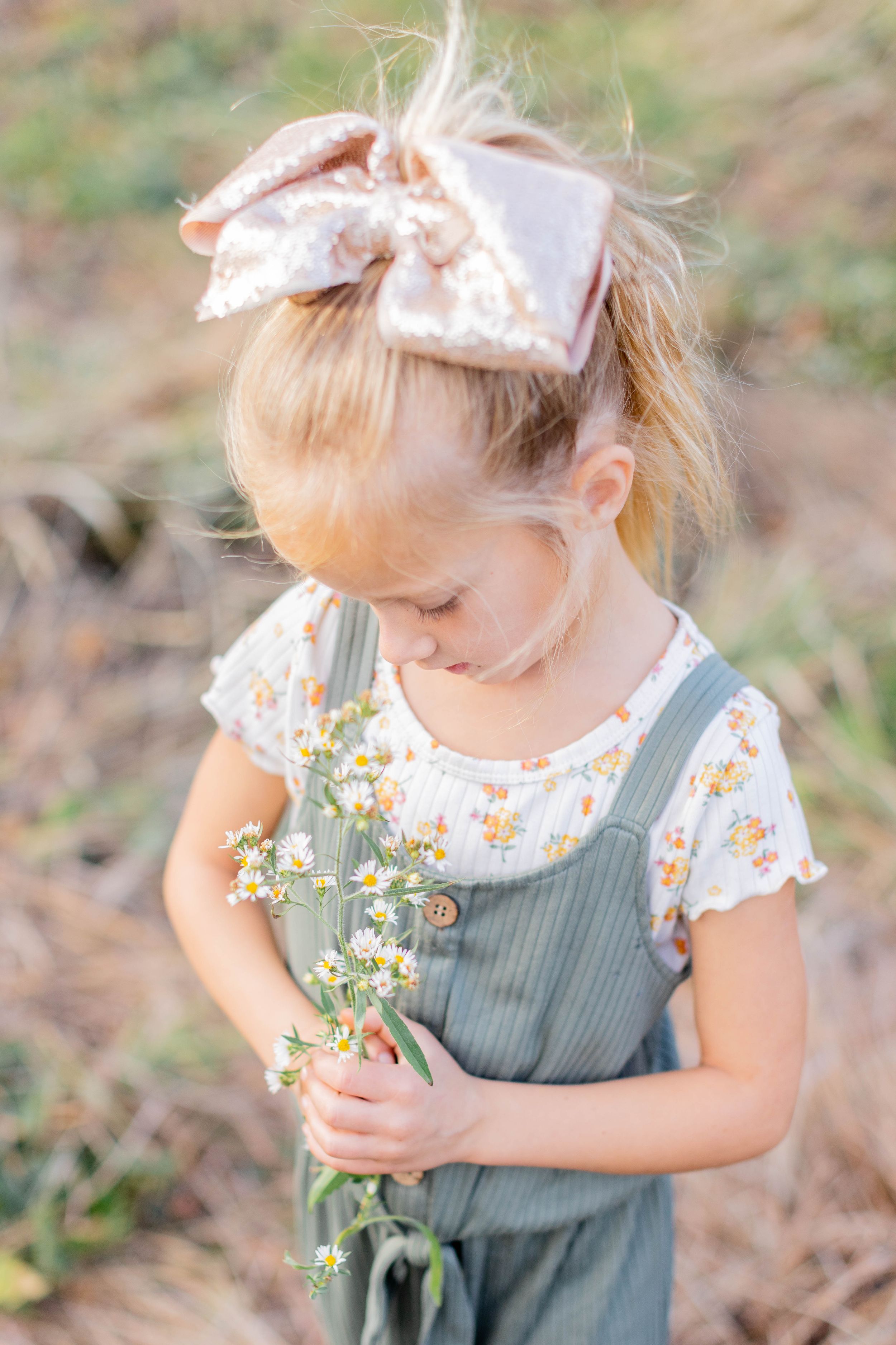 Little girl in overalls and bow holds wildflowers while looking down shyly in Springfield, MO.