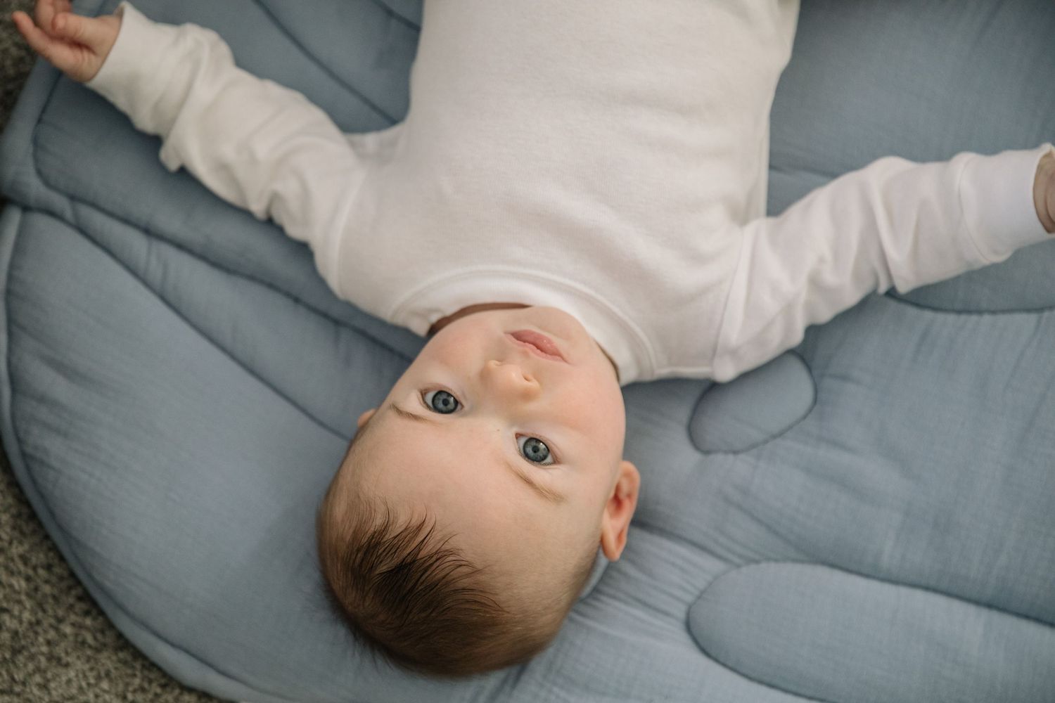 Baby in white outfit lying on blue cushion looking up at camera.