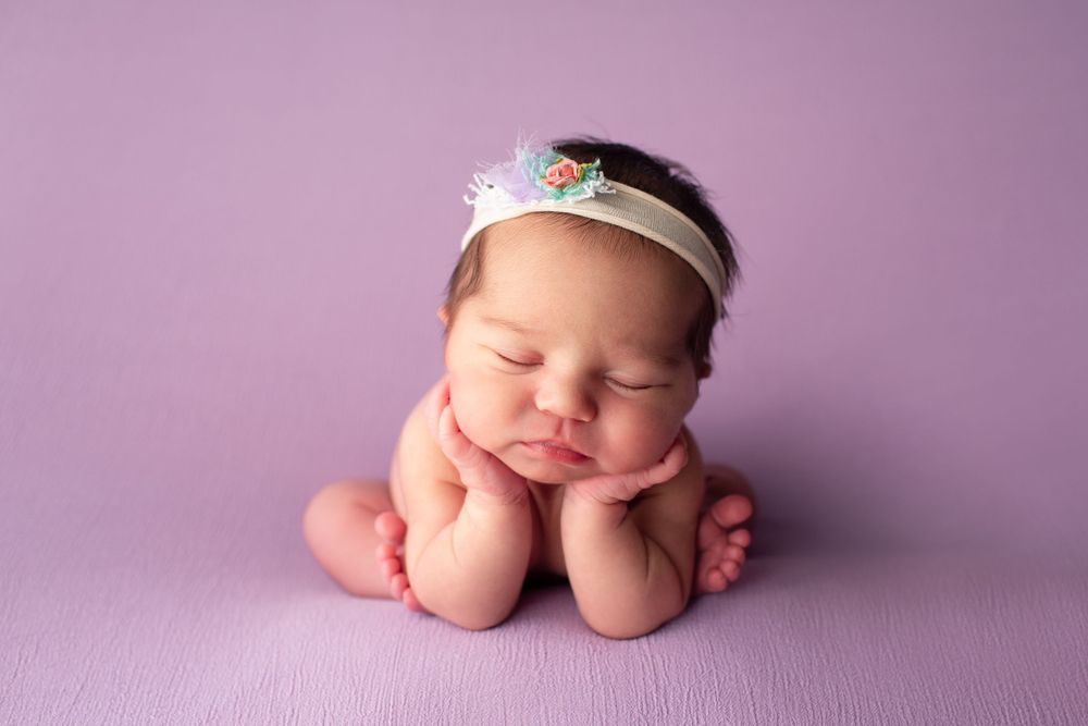 Newborn with floral headband in adorable posed position against pink backdrop.