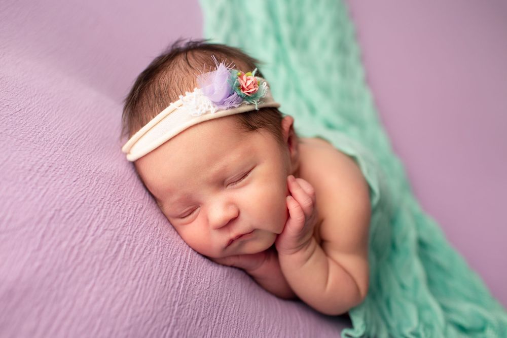 Sleeping newborn with pink floral headband against purple backdrop with mint green accent.