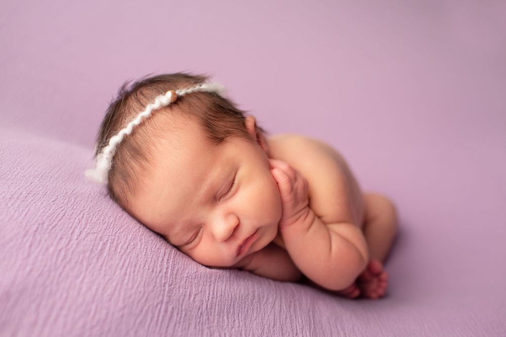 Sleeping newborn wearing delicate headband posed on soft purple backdrop during professional photo session.
