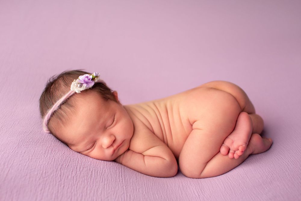 Peaceful newborn in floral headband sleeping on lavender textured backdrop during portrait session.