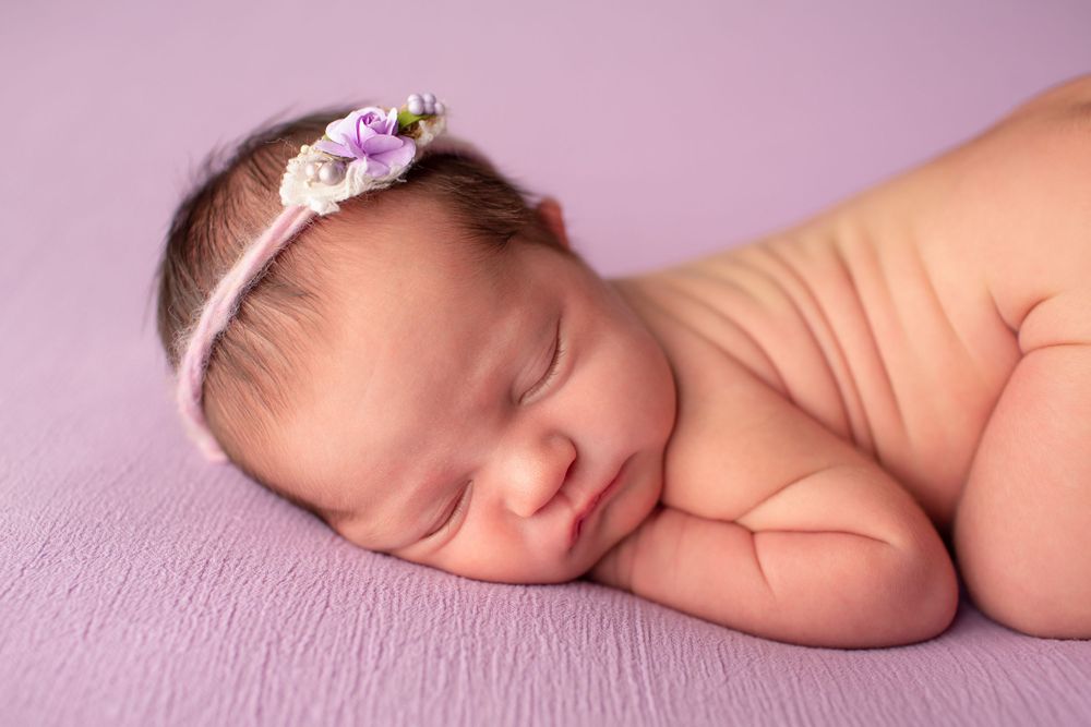 Newborn sleeping soundly on purple blanket with dainty floral headband.