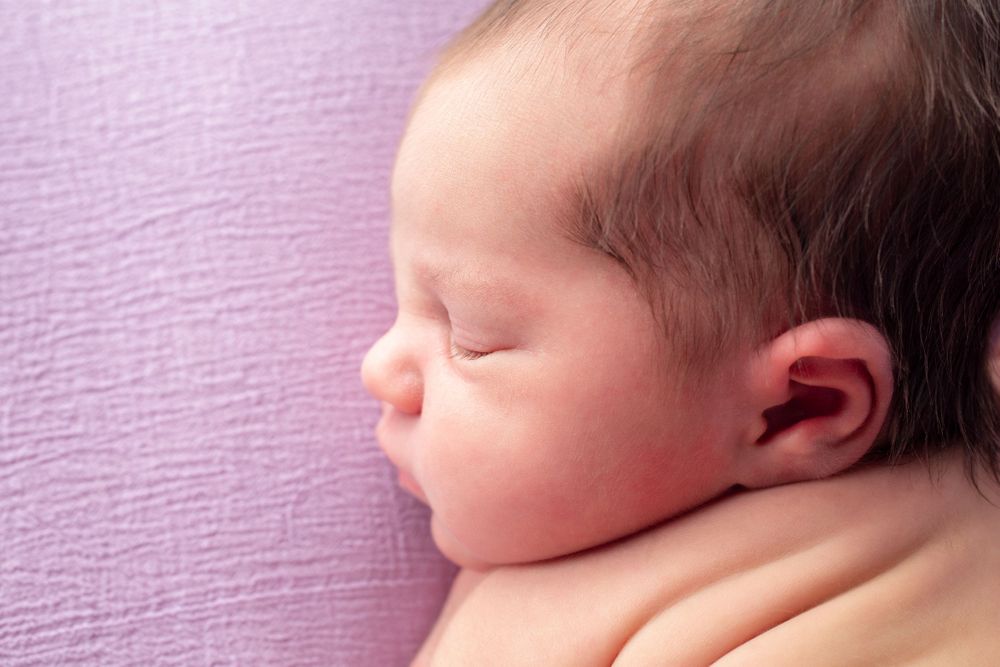 Side profile of sleeping newborn against soft purple backdrop showing delicate features.