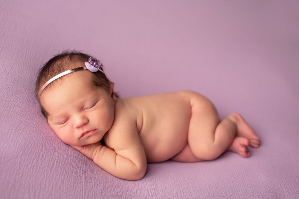 Sleeping newborn posed on purple fabric wearing delicate floral headband during professional shoot.