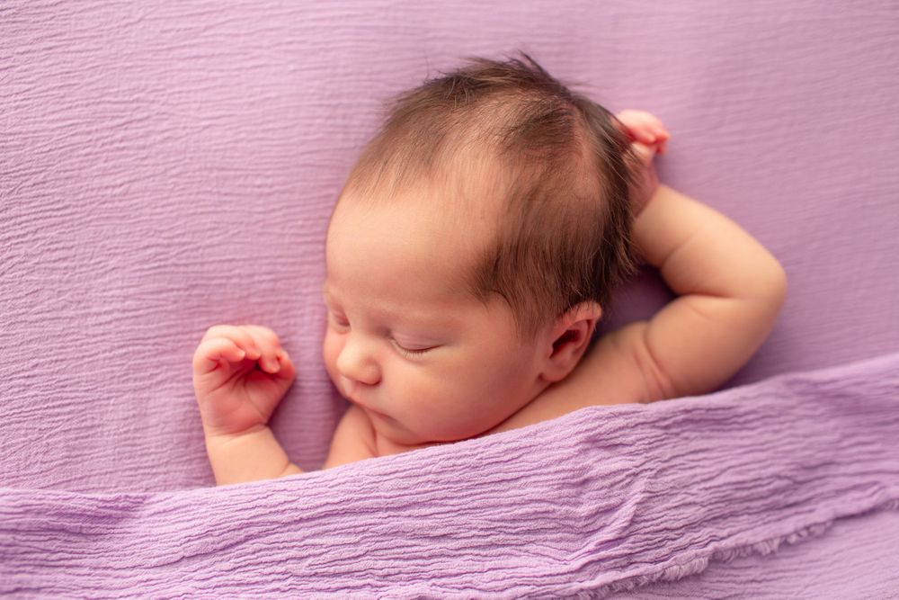 Sleeping newborn stretching on textured purple backdrop during portrait session.
