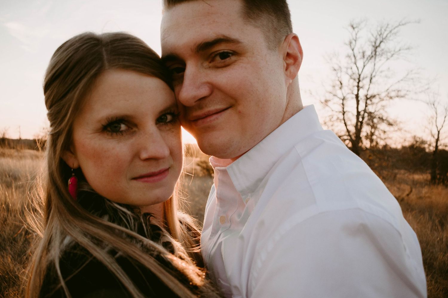 A couple shares an intimate moment at sunset in a field with tall grass and bare trees.