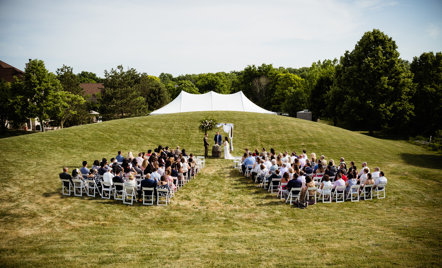 A white tent wedding ceremony setup on a grassy hilltop with rows of white chairs and guests seated.