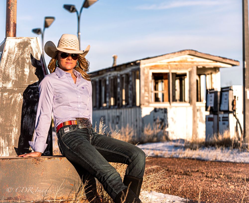 Western style portrait set against rustic building backdrop at sunset with cowboy hat and denim attire.