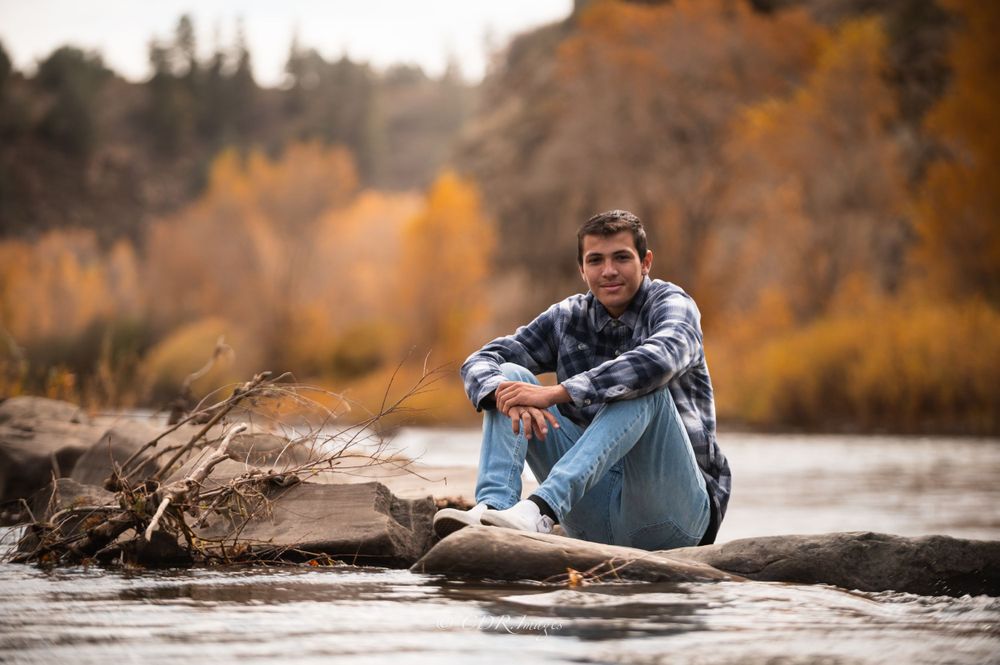 Person in casual wear sitting on rock by river with autumn colors in background.