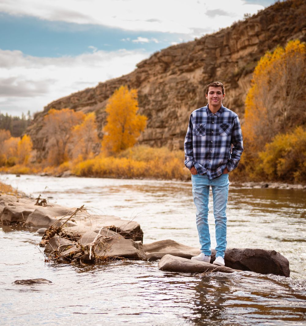 Person standing on rocks by a river with autumn foliage and mountains in the background.