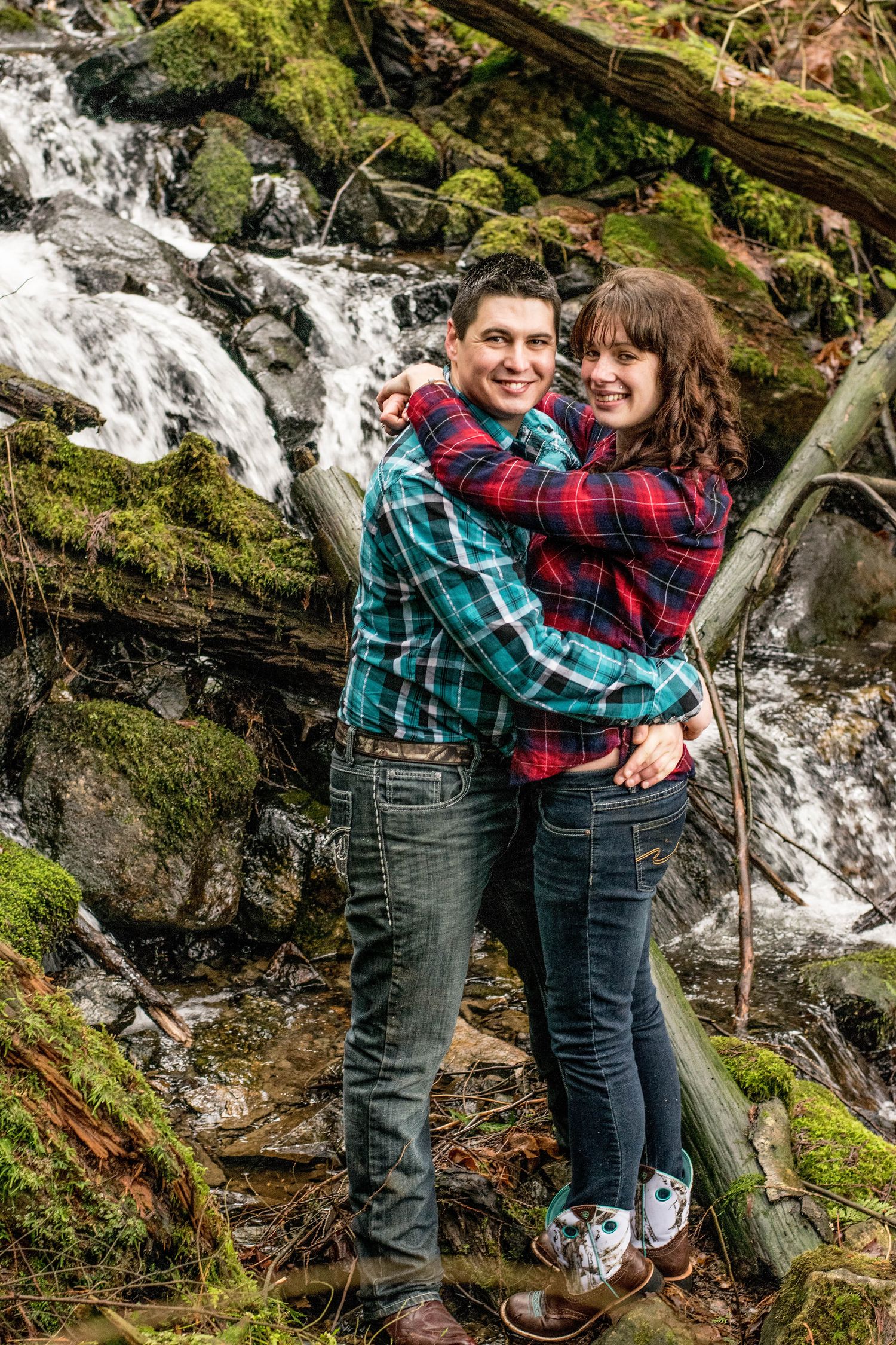 A couple in plaid shirts embrace while posing in front of a cascading waterfall.
