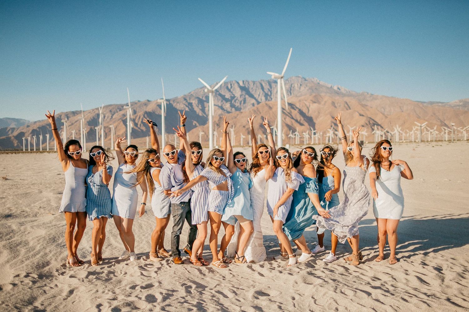 Large group poses together on sandy beach with wind turbines and mountains in background.