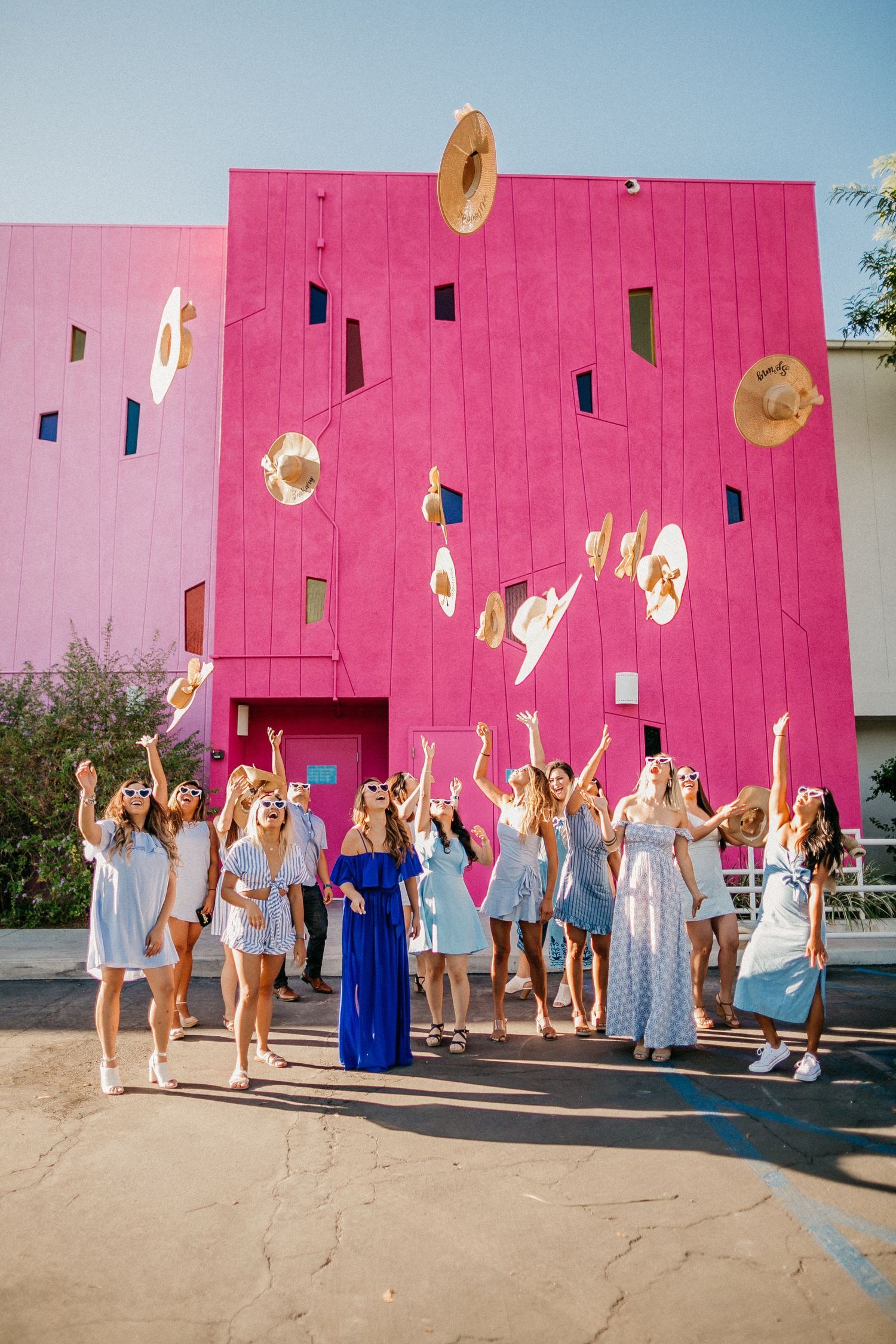 Wedding party throwing hats in front of bright pink building wall.