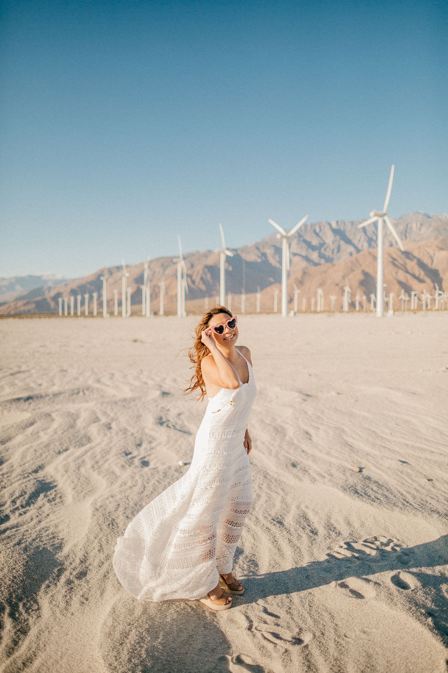 Desert photoshoot with white dress against wind turbines and mountains.