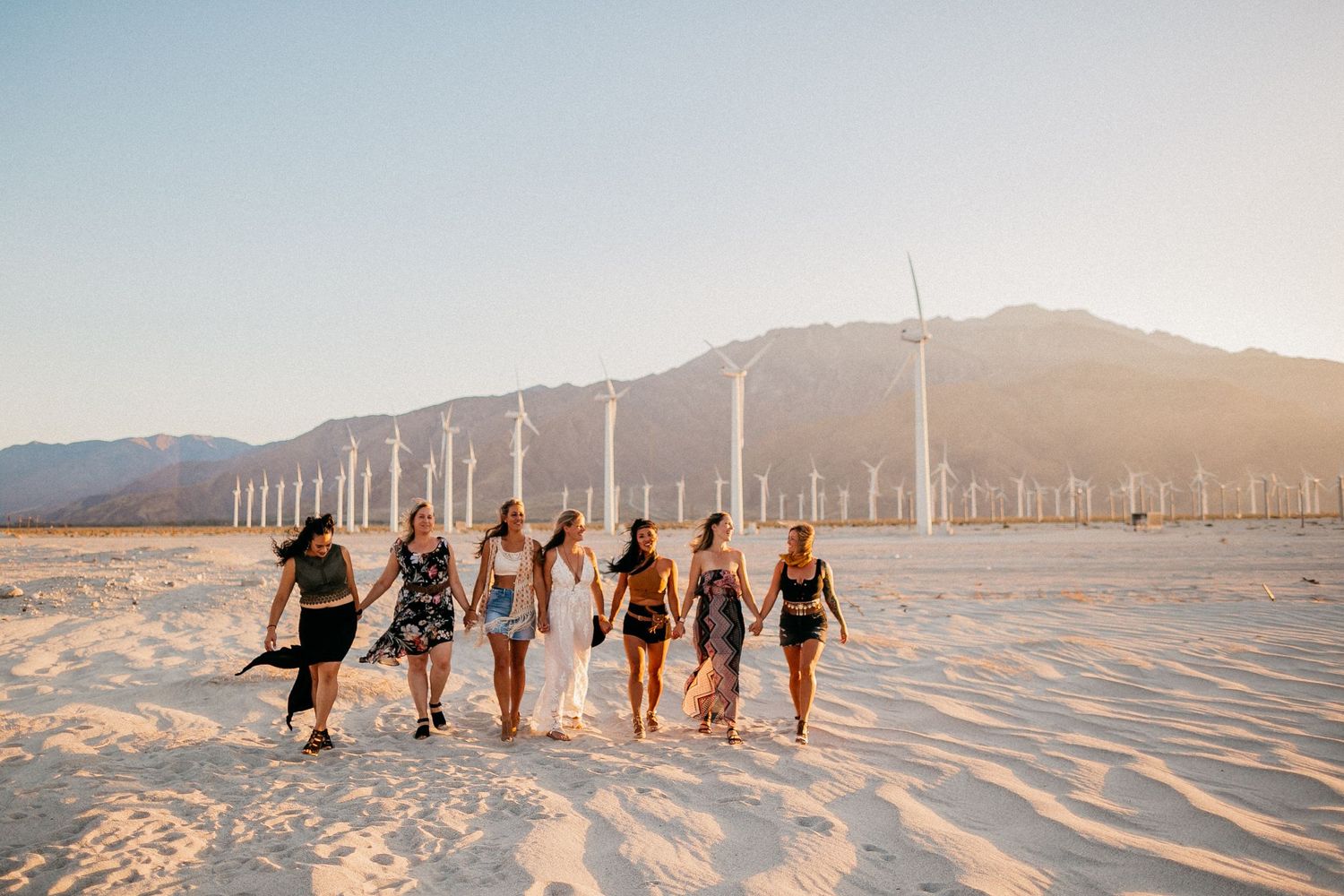 Friends walking together on sandy beach with wind turbines in background at sunset.