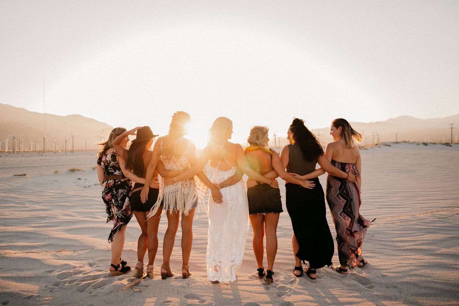 Bridal party embracing on beach during golden hour with mountains in background.