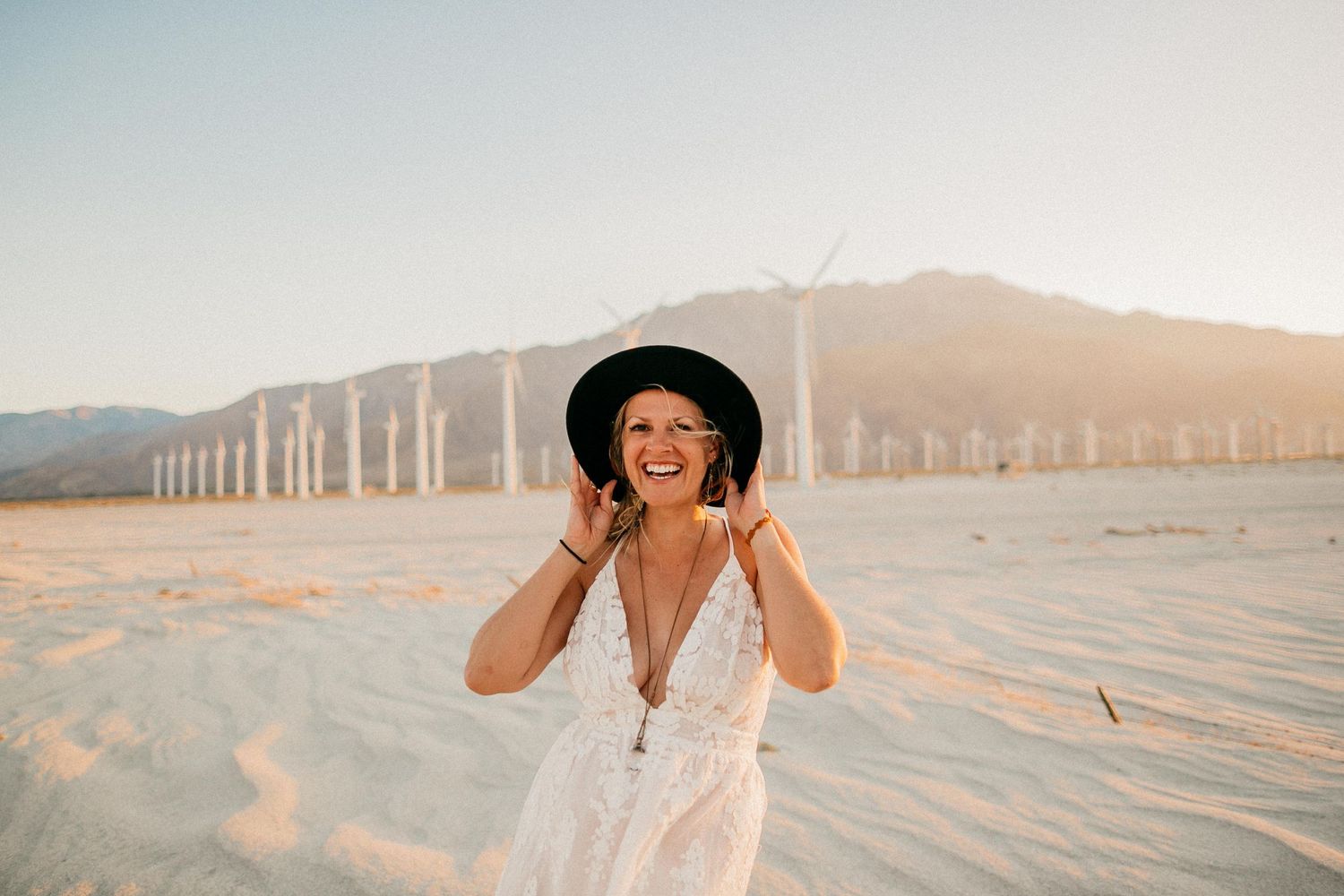 Woman in white dress and hat laughing in desert setting with wind turbines.