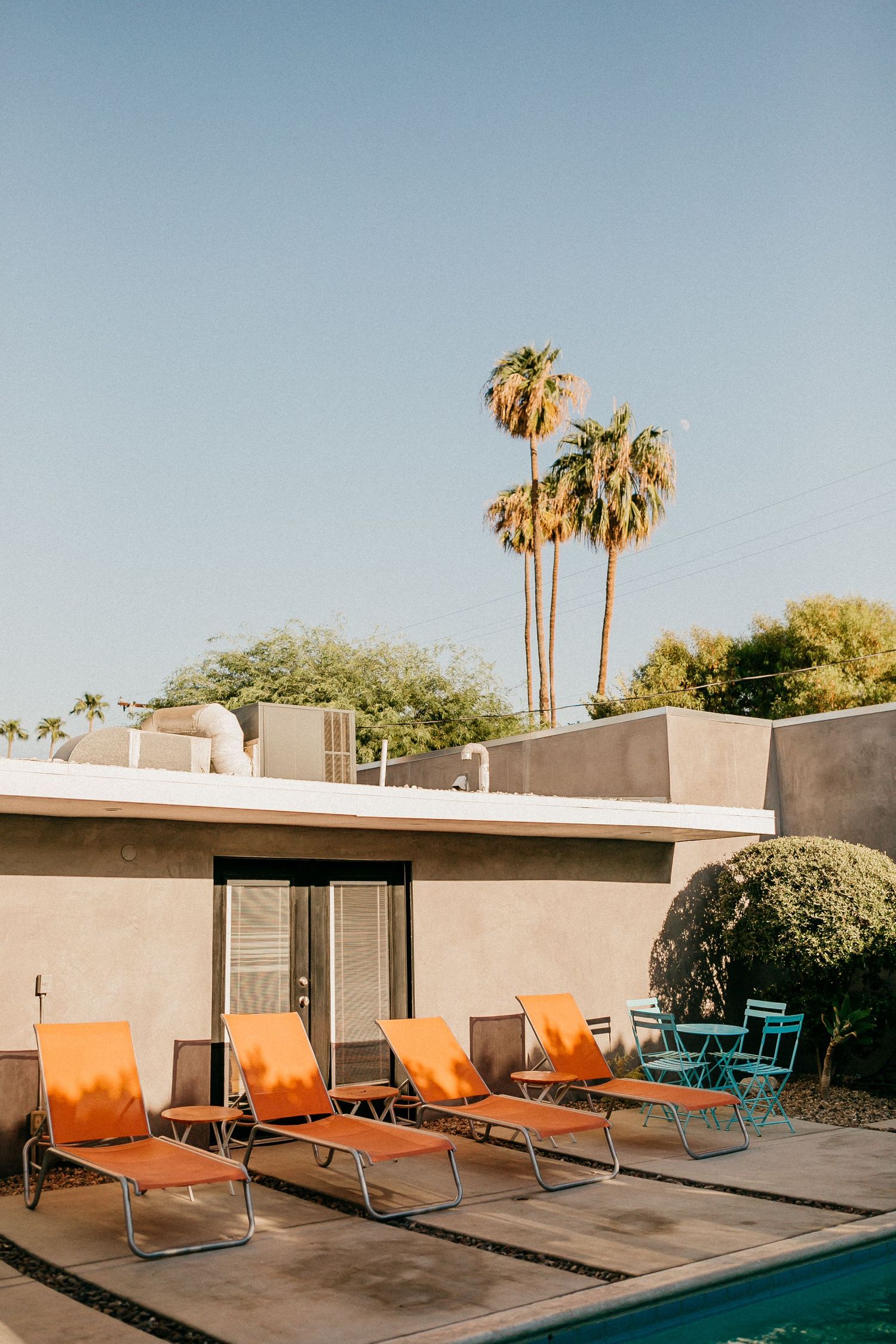 Mid-century modern poolside with orange lounge chairs and palm trees against blue sky.