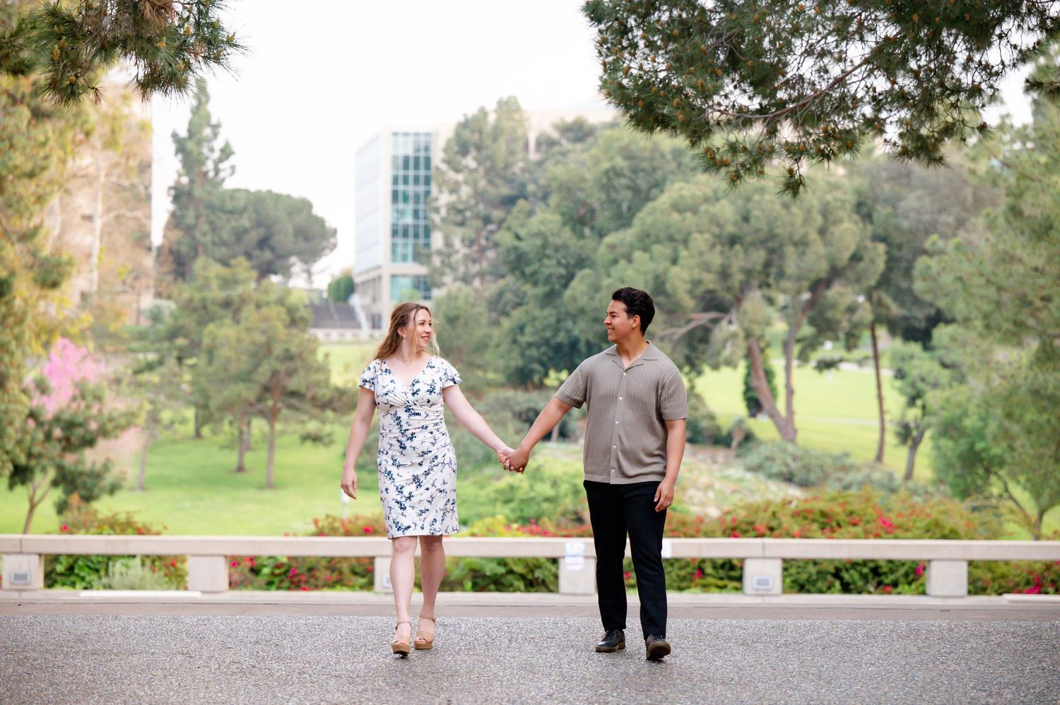 Couple holding hands while walking in park with tall buildings and trees in background.