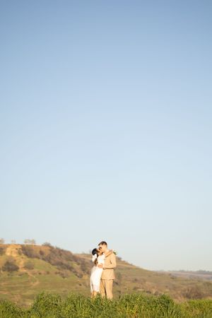 Couple sharing a romantic kiss on a lush green hillside under a clear blue sky.
