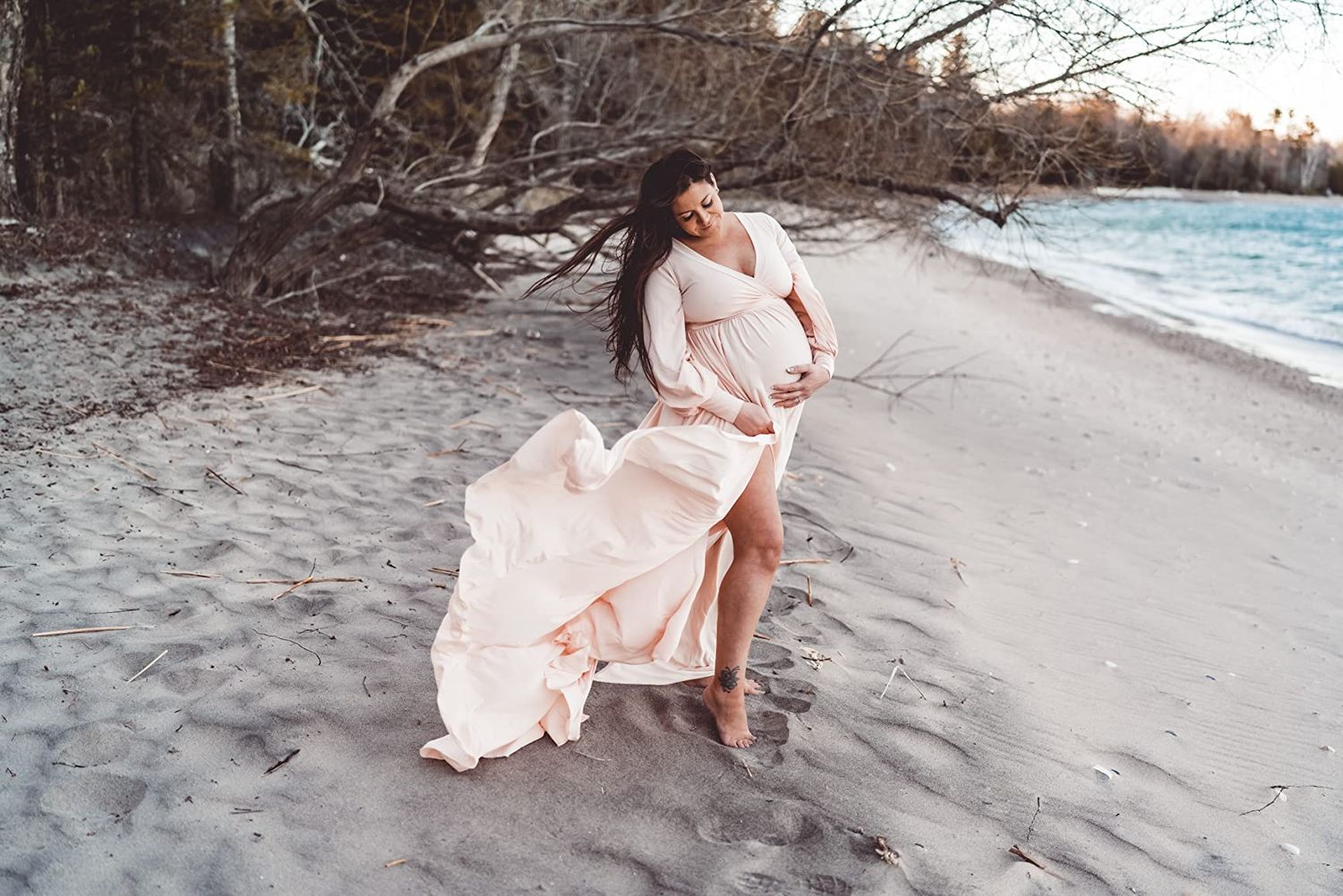 Person in flowing pink dress poses on sandy beach at sunset with water in background.