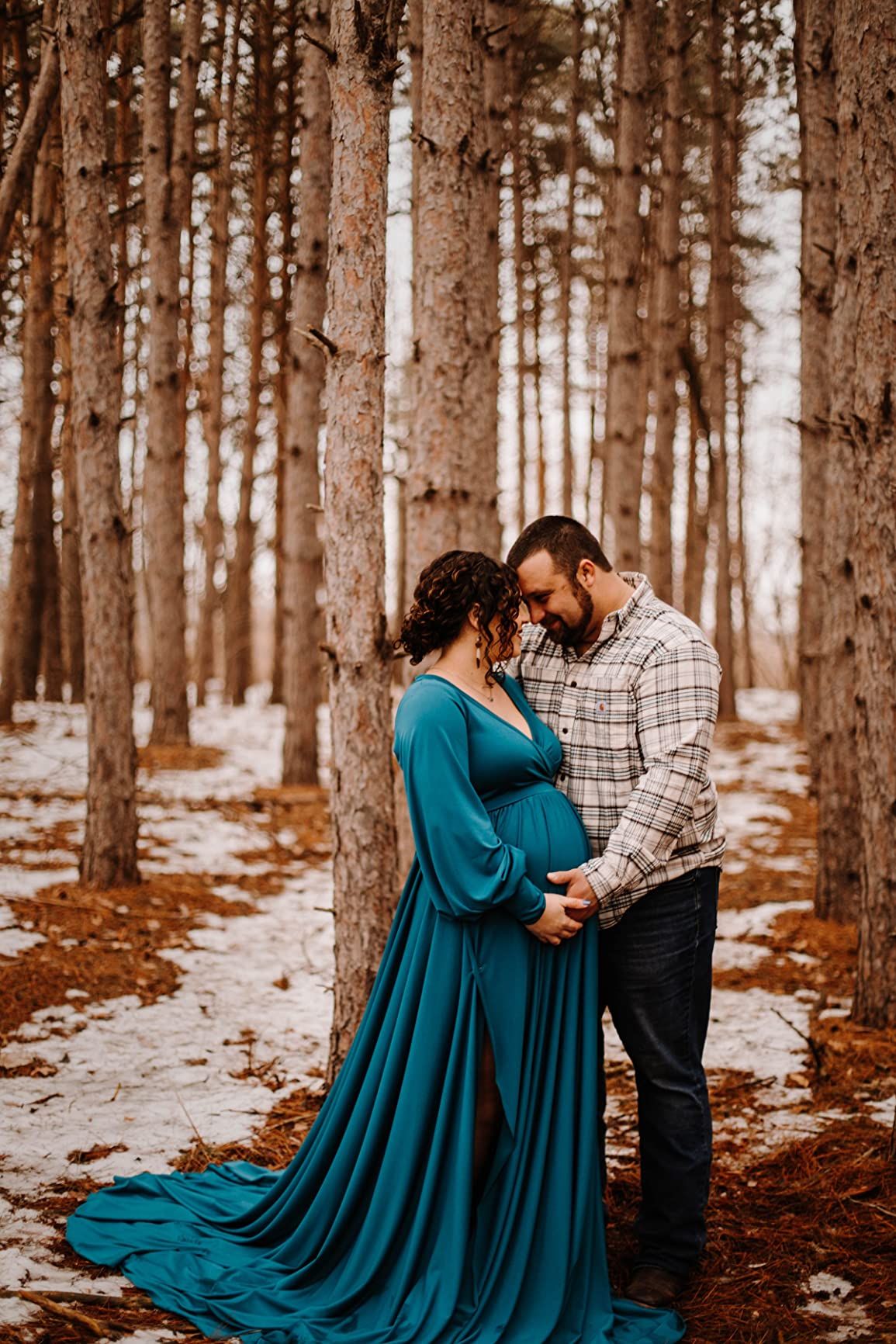A romantic maternity photo of a couple in a snowy forest with the woman wearing a teal flowing dress.