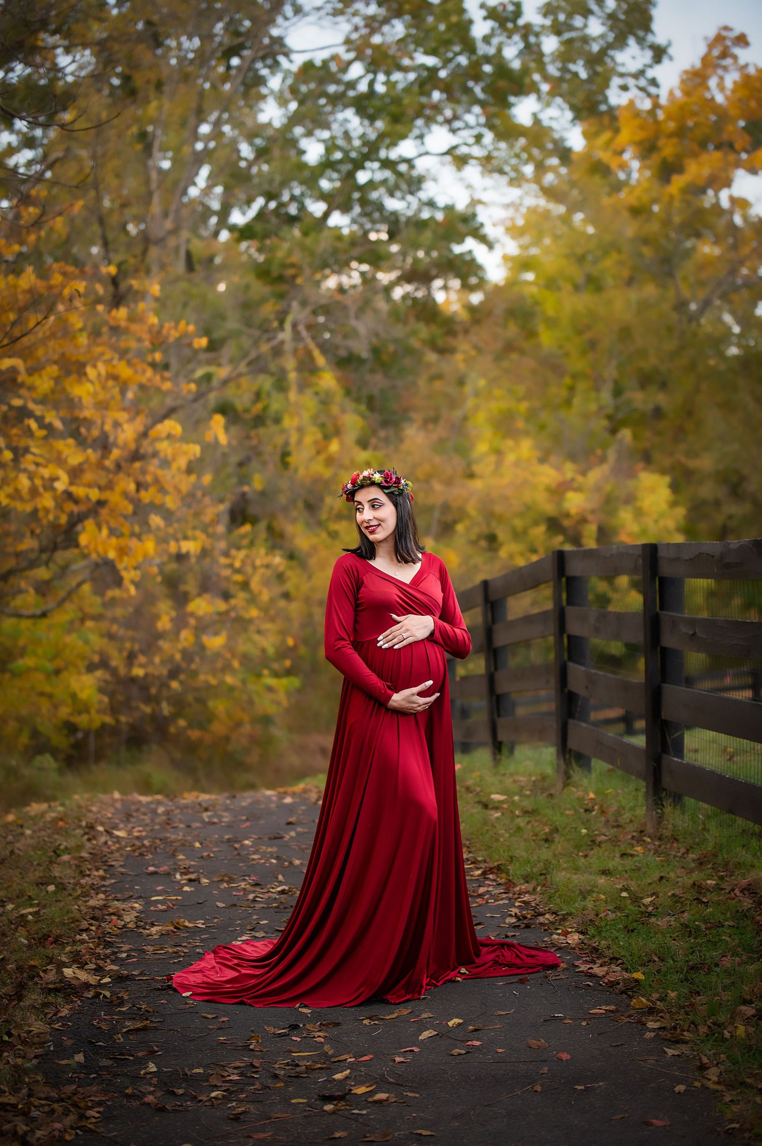 Elegant maternity photo in a red gown against autumn foliage and rustic fence.