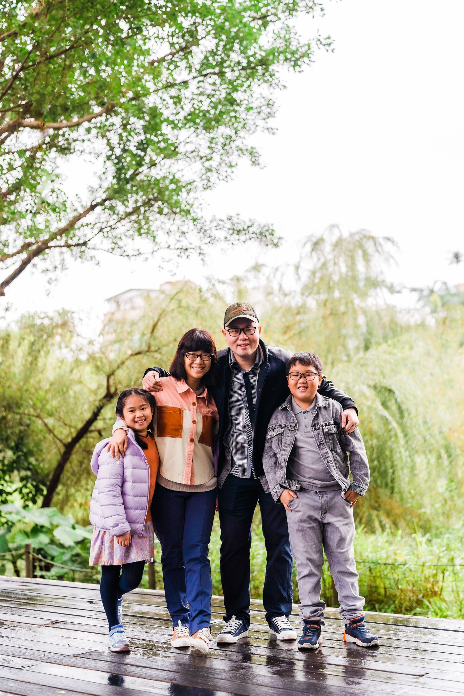 Family of four standing together on wooden deck with lush greenery in background.