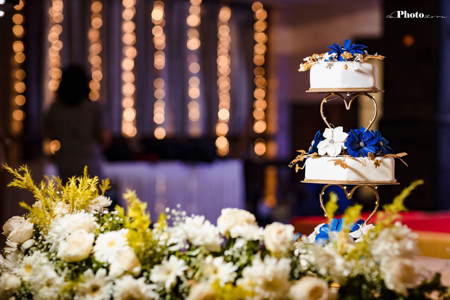 Elegant wedding cake decorated with blue flowers and string lights in background.