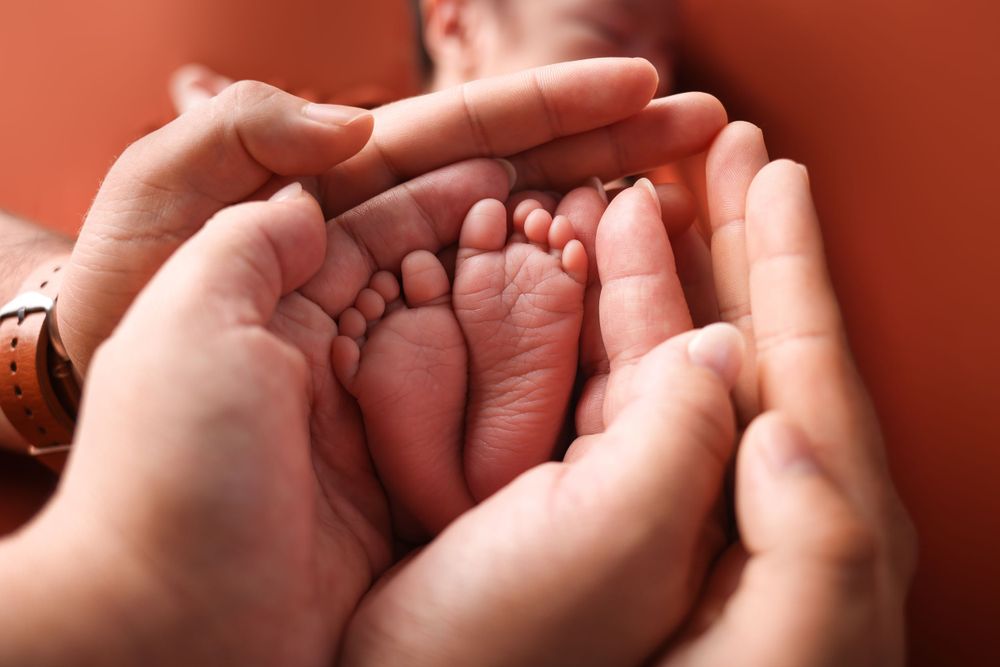 Tiny newborn baby feet being gently cradled in adult hands against a warm red background.