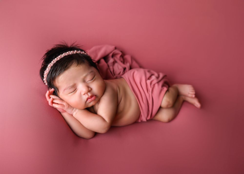 Sleeping newborn in pink wrap on matching pink background with delicate headband.
