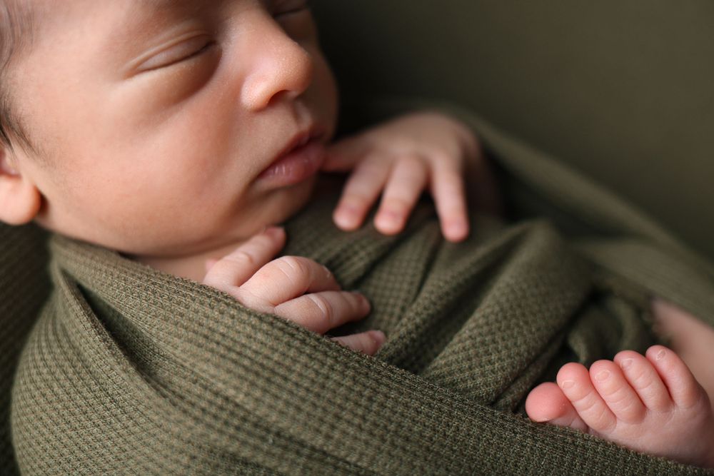 Sleeping newborn wrapped in olive green fabric with small hand visible.