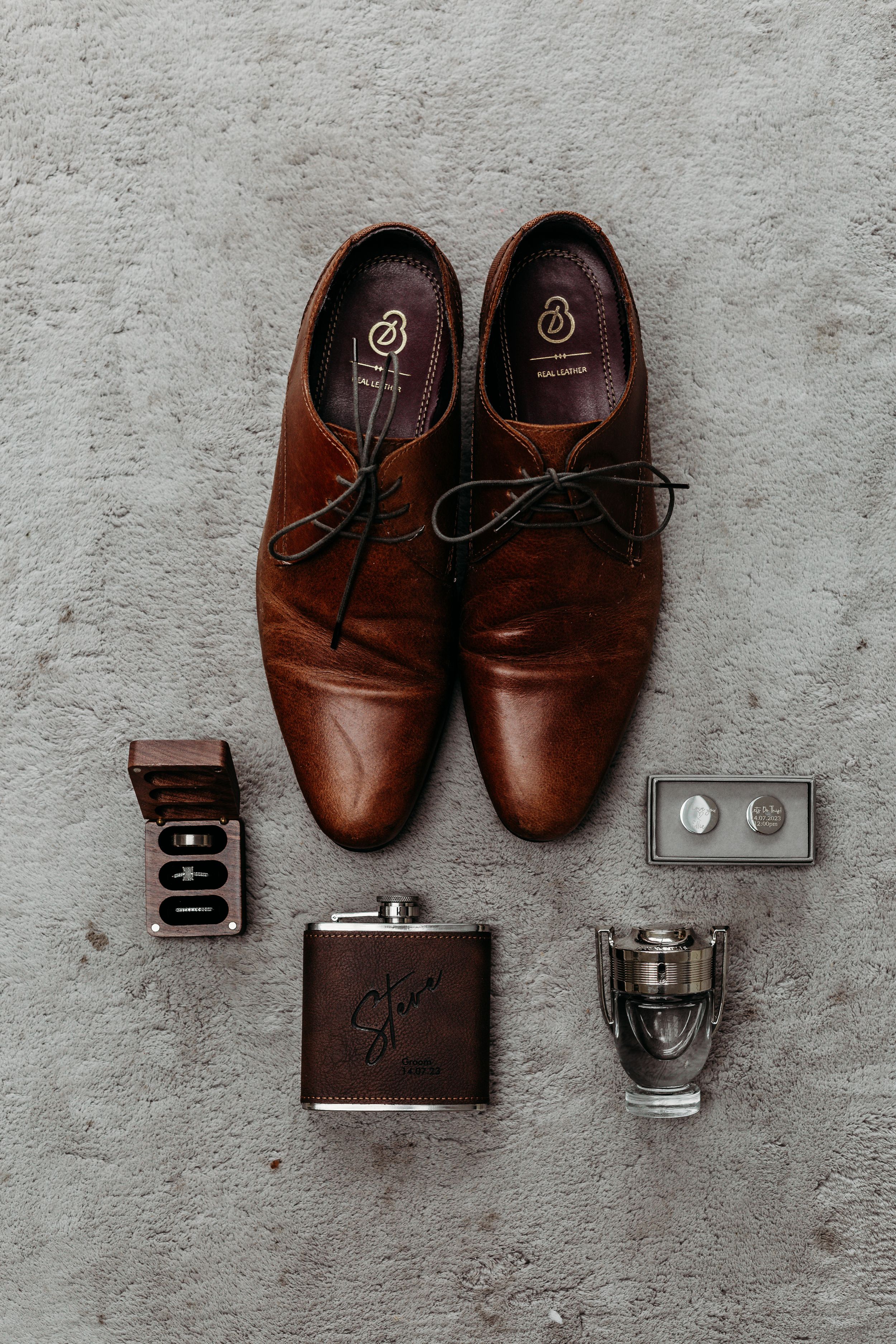 Brown leather dress shoes with wedding accessories including cufflinks, flask and rings on concrete surface.